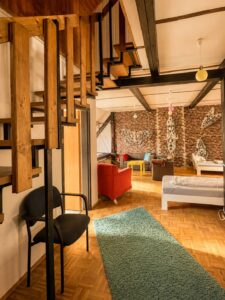 Großes Mehrbettzimmer im Homeplanet Hostel Leipzig Connewitz mit Betten, Sitzgelegenheiten, Teppich, Treppe zu einer Schlafebene und Gemeinschaftsbereich. Schlafplätze und Aufenthaltsbereich befinden sich in einem gemeinsamen Raum.
