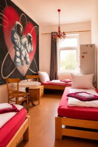 Mehrbettzimmer im Homeplanet Hostel Leipzig Connewitz mit mehreren Einzelbetten, Tisch, Stühlen, Fenster und Wandgestaltung. Die Schlafplätze sind im Raum verteilt angeordnet.