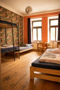 Mehrbettzimmer im Homeplanet Hostel Leipzig Connewitz mit mehreren Betten, Etagenbett, Tisch, Stühlen und Fenstern. Die Schlafplätze sind im Raum verteilt angeordnet.