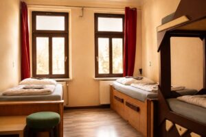 Mehrbettzimmer im Homeplanet Hostel Leipzig Connewitz mit Einzelbetten, Etagenbett, Fenstern, Sitzhocker und Stauraum unter den Betten. Die Schlafplätze sind entlang der Wände angeordnet.