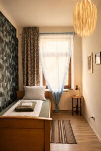 Einzelzimmer im Homeplanet Hostel Leipzig Connewitz mit Einzelbett, Fenster, Nachttisch, Leselampe und Teppich. Das Bett befindet sich entlang der Wand.