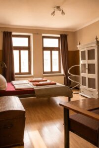 Doppelzimmer im Homeplanet Hostel Leipzig Connewitz mit Doppelbett, Kleiderschrank, Sitzgelegenheit, Truhe und zwei Fenstern. Das Bett befindet sich mittig im Raum.